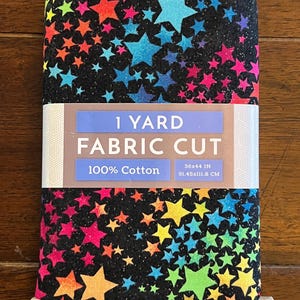 May include: A fabric cut featuring a vibrant star pattern in shades of blue, pink, yellow, green, and orange on a black background. The fabric is 36x44 inches and made of 100% cotton. The packaging reads "1 Yard Fabric Cut".