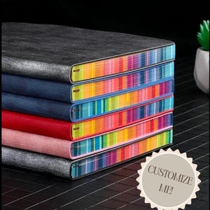 May include: A stack of colorful notebooks with striped covers. The top notebook is black with a textured cover. The notebooks are stacked on a black surface. The text "CUSTOMIZE ME!" is on a white sticker.