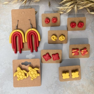 Könnte beinhalten: Sortiment handgefertigter Ohrringe aus Polymer Clay in Rot und Gelb. Die Ohrringe umfassen ein Paar Regenbogenbögen, ein Paar Schildkröten, ein Paar Kissen, ein Paar Knoten, ein Paar Wirbel und ein Paar Quadrate.