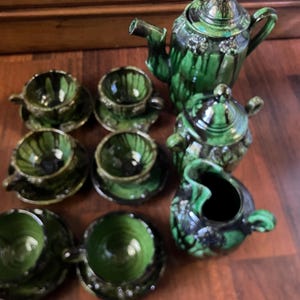 Peut inclure: Un service à thé vintage en céramique verte, comprenant une théière, un sucrier, une crémière et six tasses avec soucoupes. L'ensemble a une finition brillante avec des accents vert foncé et noirs. Le service à thé est présenté sur une surface en bois.