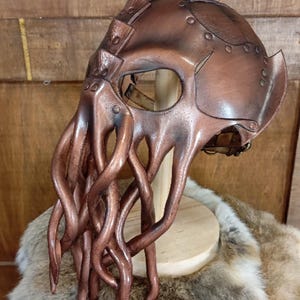 May include: A brown leather mask featuring a smooth, rounded headpiece and multiple long, dangling tentacles extending from the front. The mask is constructed with visible metal rivets and has a large, singular eye opening for visibility.