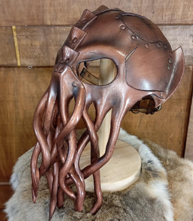 May include: A brown leather mask featuring a smooth, rounded headpiece and multiple long, dangling tentacles extending from the front. The mask is constructed with visible metal rivets and has a large, singular eye opening for visibility.