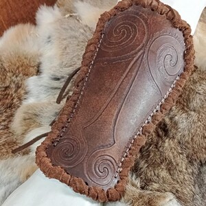 May include: A brown leather bracer featuring embossed spiral patterns and a thick, braided leather edge. The piece is constructed with visible stitching along the perimeter and includes thin leather lacing cords extending from the side.