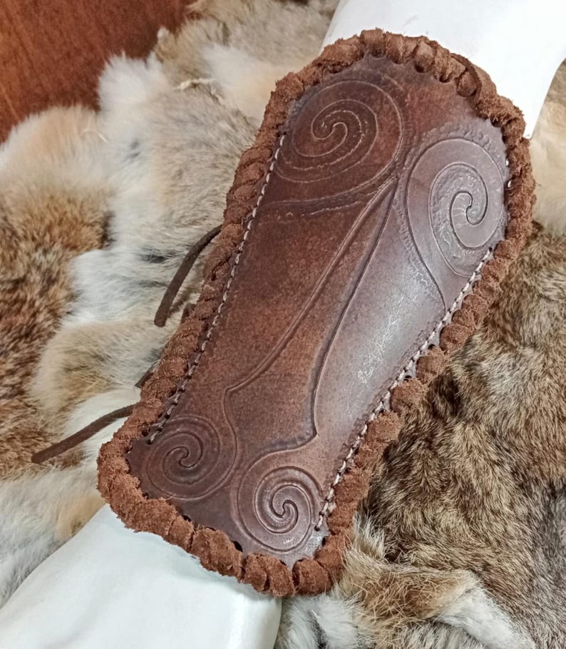 May include: A brown leather bracer featuring embossed spiral patterns and a thick, braided leather edge. The piece is constructed with visible stitching along the perimeter and includes thin leather lacing cords extending from the side.