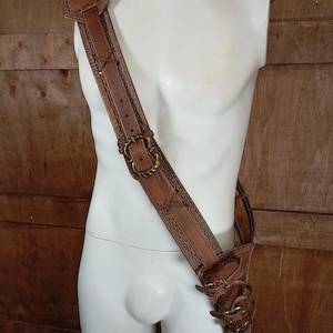 May include: A tan leather shoulder strap and holster assembly featuring intricate embossed border patterns, antique brass-toned metal buckles, and multiple adjustable leather straps secured with metal hardware, worn diagonally across the torso.