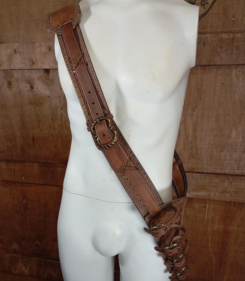 May include: A tan leather shoulder strap and holster assembly featuring intricate embossed border patterns, antique brass-toned metal buckles, and multiple adjustable leather straps secured with metal hardware, worn diagonally across the torso.