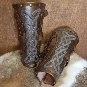 May include: A pair of brown leather arm guards with silver Celtic knot designs and metal rivets. The guards have adjustable straps.