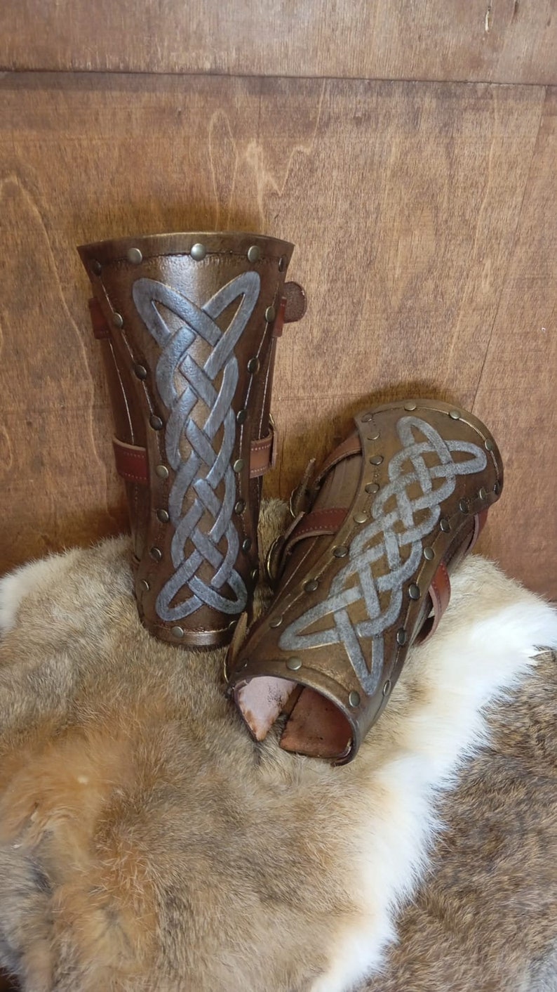 May include: A pair of brown leather arm guards with silver Celtic knot designs and metal rivets. The guards have adjustable straps.