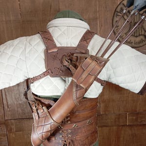 May include: A brown leather archery quiver and arm guard set. The quiver is attached to a brown leather harness and a quilted white jacket. The set includes several arrows with black fletching. The background is a wooden wall.