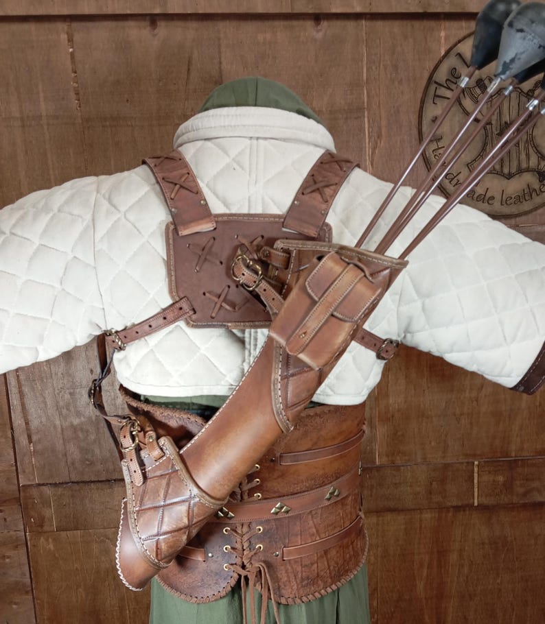 May include: A brown leather archery quiver and arm guard set. The quiver is attached to a brown leather harness and a quilted white jacket. The set includes several arrows with black fletching. The background is a wooden wall.