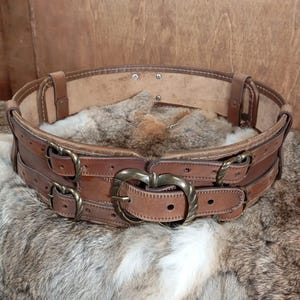 May include: A wide, brown leather belt with multiple straps and brass buckles. The belt has a decorative, heart-shaped buckle in the center. The belt is shown on a fur surface.
