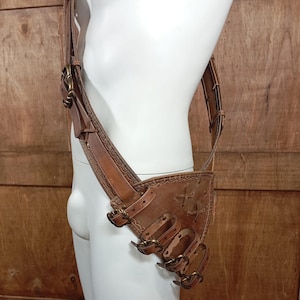 May include: A tan leather harness featuring a wide shoulder strap and a triangular hip pouch. The design includes multiple adjustable straps with brass-toned metal buckles, decorative stitching along the edges, and crisscross lacing details.