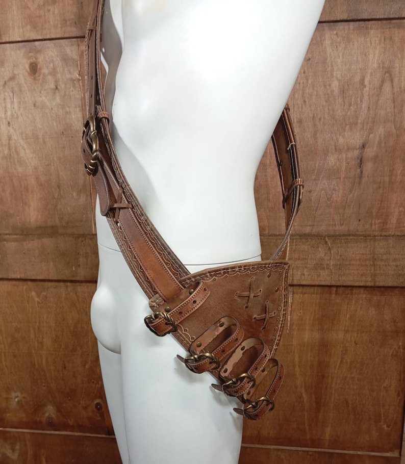 May include: A tan leather harness featuring a wide shoulder strap and a triangular hip pouch. The design includes multiple adjustable straps with brass-toned metal buckles, decorative stitching along the edges, and crisscross lacing details.