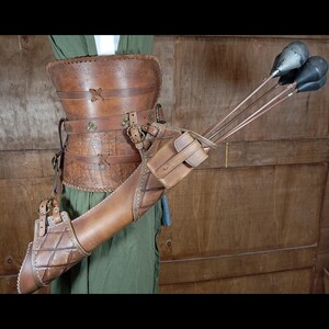 May include: A brown leather archery quiver and belt set. The quiver is cylindrical with a small pouch and holds several arrows. The belt has decorative stitching and metal accents. The set is designed to be worn around the waist.