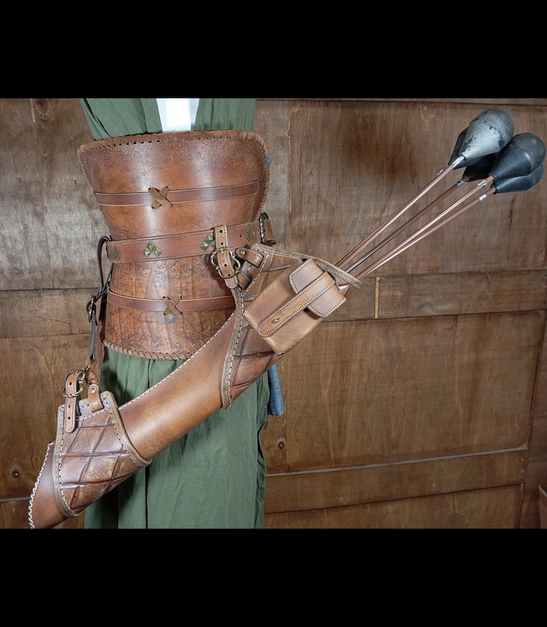 May include: A brown leather archery quiver and belt set. The quiver is cylindrical with a small pouch and holds several arrows. The belt has decorative stitching and metal accents. The set is designed to be worn around the waist.