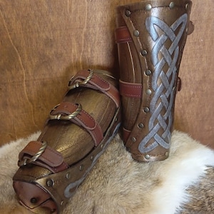 May include: Two brown leather arm guards with metal accents and a Celtic knot design. The arm guards are on a brown and white fur surface.