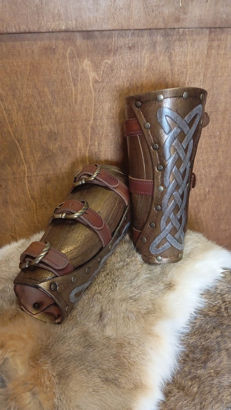 May include: Two brown leather arm guards with metal accents and a Celtic knot design. The arm guards are on a brown and white fur surface.