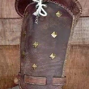 May include: A brown leather arm guard with decorative stitching and metal studs. The guard has a strap with a buckle.
