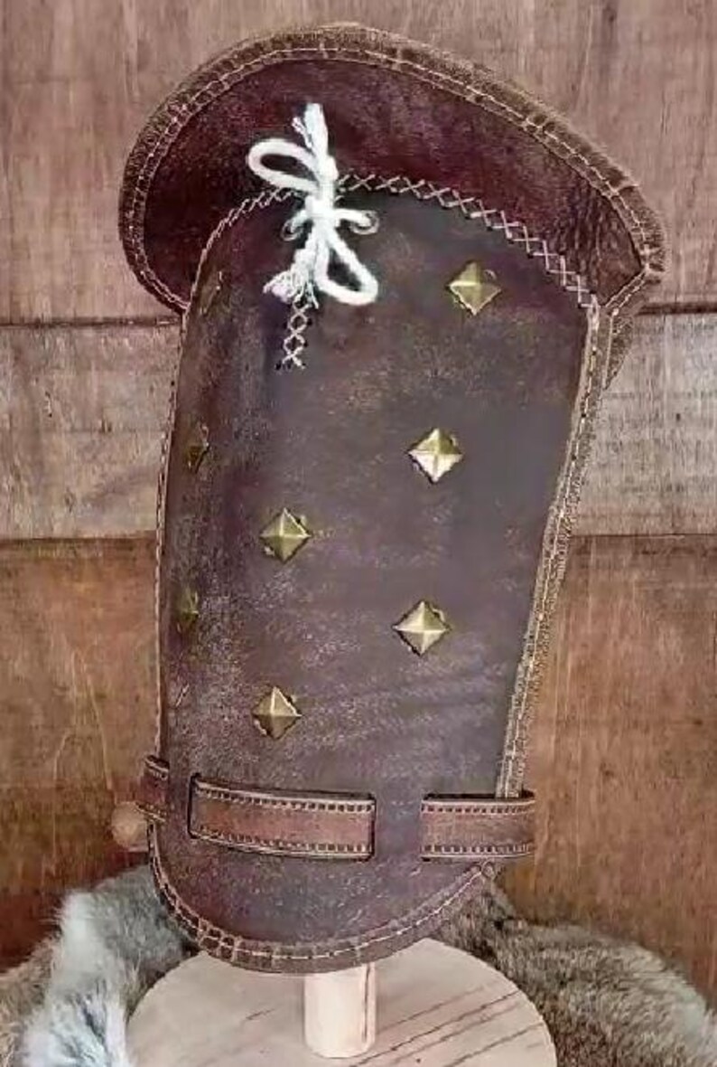 May include: A brown leather arm guard with decorative stitching and metal studs. The guard has a strap with a buckle.