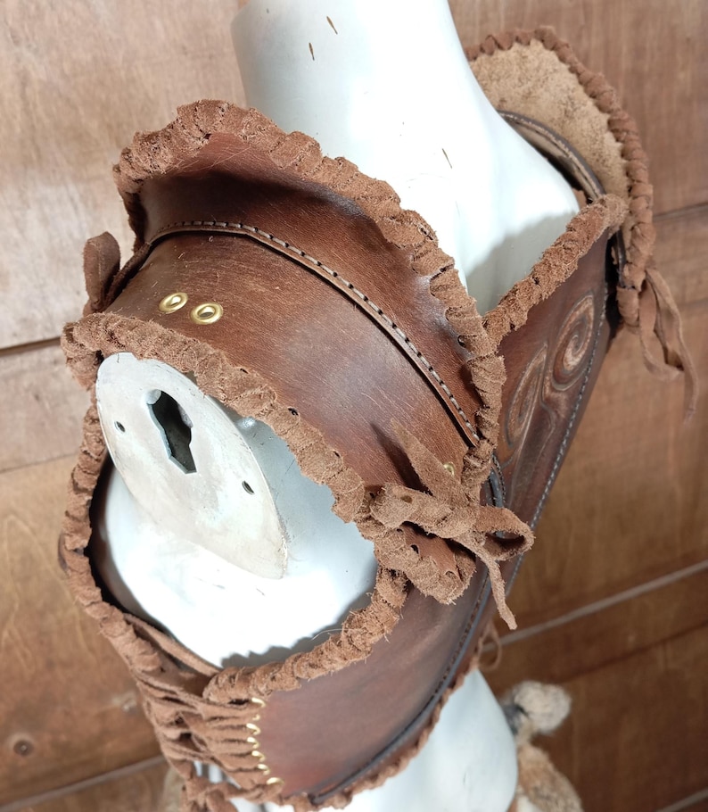 May include: A brown leather chest piece featuring decorative embossed swirl patterns, finished with a braided suede trim along the edges. The garment includes gold-toned metal eyelets and a lace-up front closure, secured on a white mannequin torso.