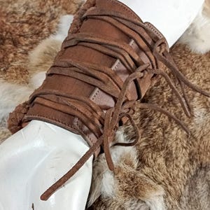May include: A brown leather wrist cuff featuring a series of horizontal leather cord laces that wrap around the circumference. The cuff has a smooth leather exterior and is lined with a textured, fuzzy brown material along the edges.