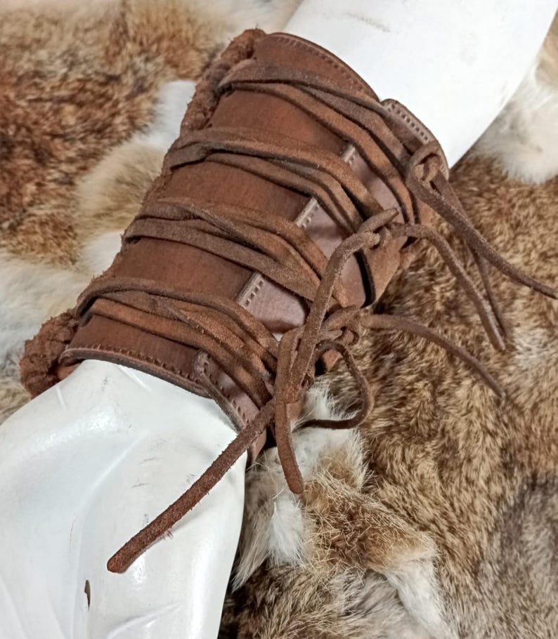 May include: A brown leather wrist cuff featuring a series of horizontal leather cord laces that wrap around the circumference. The cuff has a smooth leather exterior and is lined with a textured, fuzzy brown material along the edges.