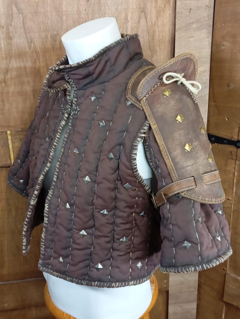 May include: A brown quilted vest with a leather shoulder pad. The vest has a high collar and is decorated with metal studs. The shoulder pad is also decorated with metal studs and has a strap that goes around the arm.
