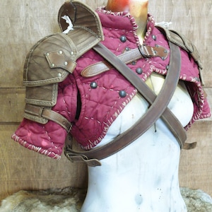 Puede incluir: Un conjunto de hombreras de estilo medieval en burdeos y marr&oacute;n. La armadura presenta tela burdeos acolchada, detalles de cuero marr&oacute;n y tachuelas met&aacute;licas. El conjunto incluye hombreras y una pieza para el pecho con correas de cuero.