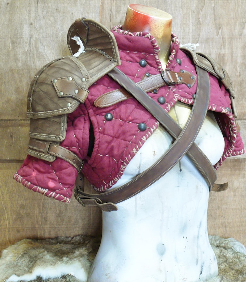 Puede incluir: Un conjunto de hombreras de estilo medieval en burdeos y marr&oacute;n. La armadura presenta tela burdeos acolchada, detalles de cuero marr&oacute;n y tachuelas met&aacute;licas. El conjunto incluye hombreras y una pieza para el pecho con correas de cuero.