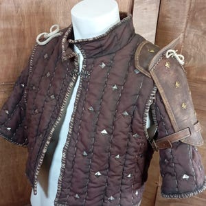 May include: A brown quilted armour vest with silver studs and a brown leather shoulder piece with straps.