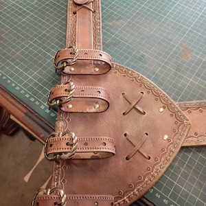 May include: A brown leather belt featuring a wide, triangular side panel with four adjustable straps secured by antique brass-toned metal buckles. The leather is decorated with embossed border patterns, cross-lacing details, and small metal rivets.