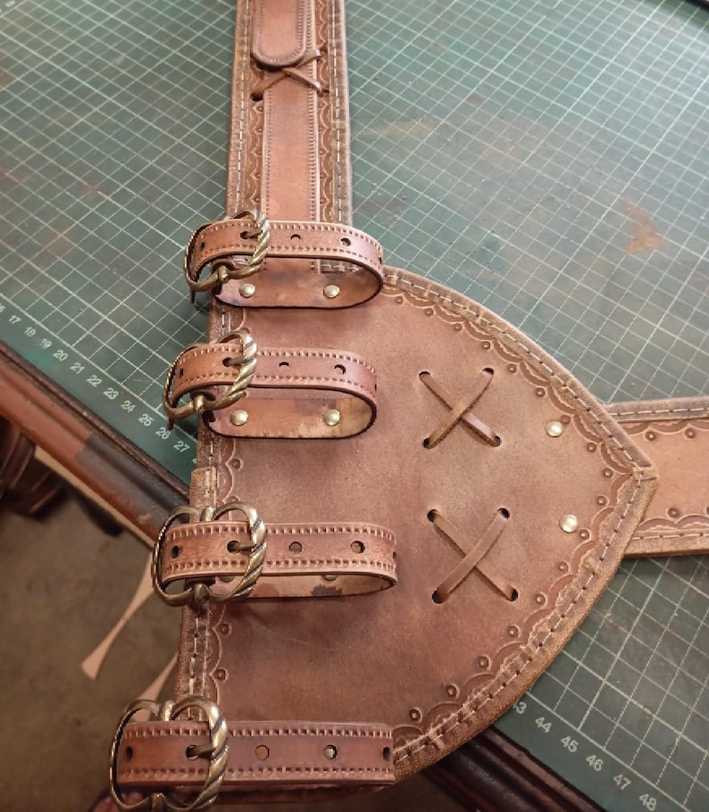 May include: A brown leather belt featuring a wide, triangular side panel with four adjustable straps secured by antique brass-toned metal buckles. The leather is decorated with embossed border patterns, cross-lacing details, and small metal rivets.