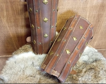 May include: Two brown leather arm guards with brass-colored studs and rivets. The arm guards are cylindrical with straps and buckles. The text "DIGITAL DOWNLOAD" and "PATTERN & INSTRUCTIONS INCLUDED" is at the bottom of the image.