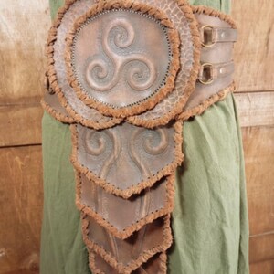 May include: A brown leather belt featuring a large circular centerpiece with a triskelion symbol, surrounded by layered, scale-like panels. The edges of the leather pieces are finished with a textured, frayed brown trim and secured with metal rings.