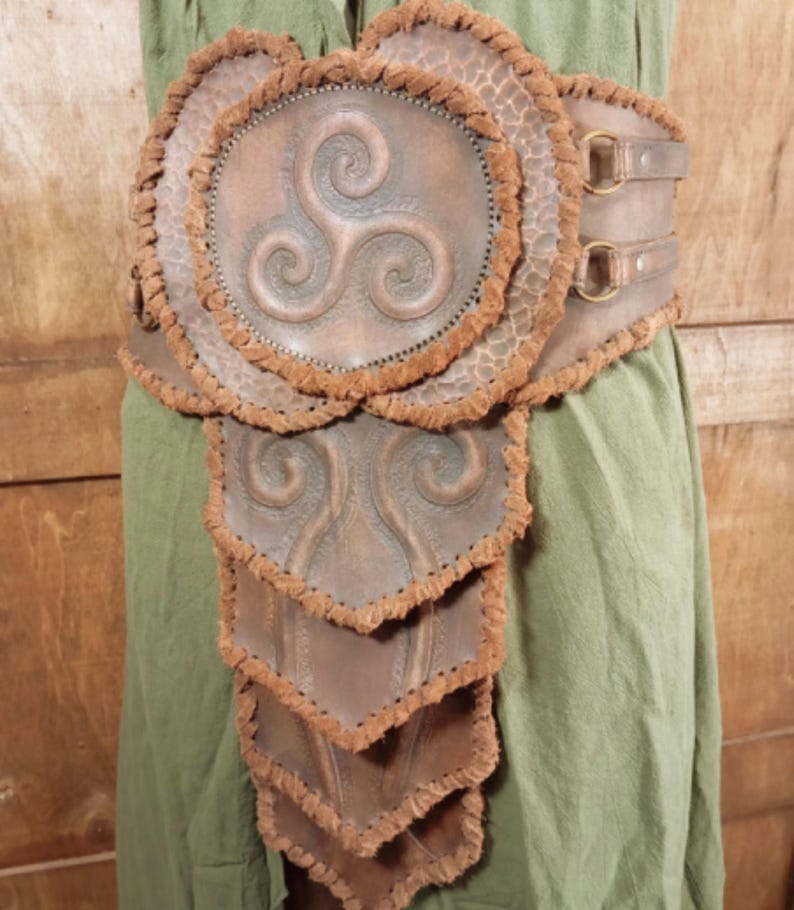 May include: A brown leather belt featuring a large circular centerpiece with a triskelion symbol, surrounded by layered, scale-like panels. The edges of the leather pieces are finished with a textured, frayed brown trim and secured with metal rings.