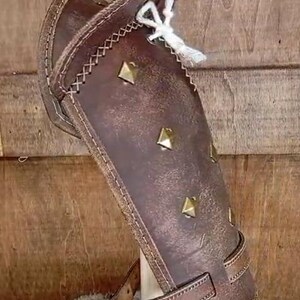 May include: Brown leather arm guard with decorative stitching and metal studs. The guard has a strap with a buckle for fastening.