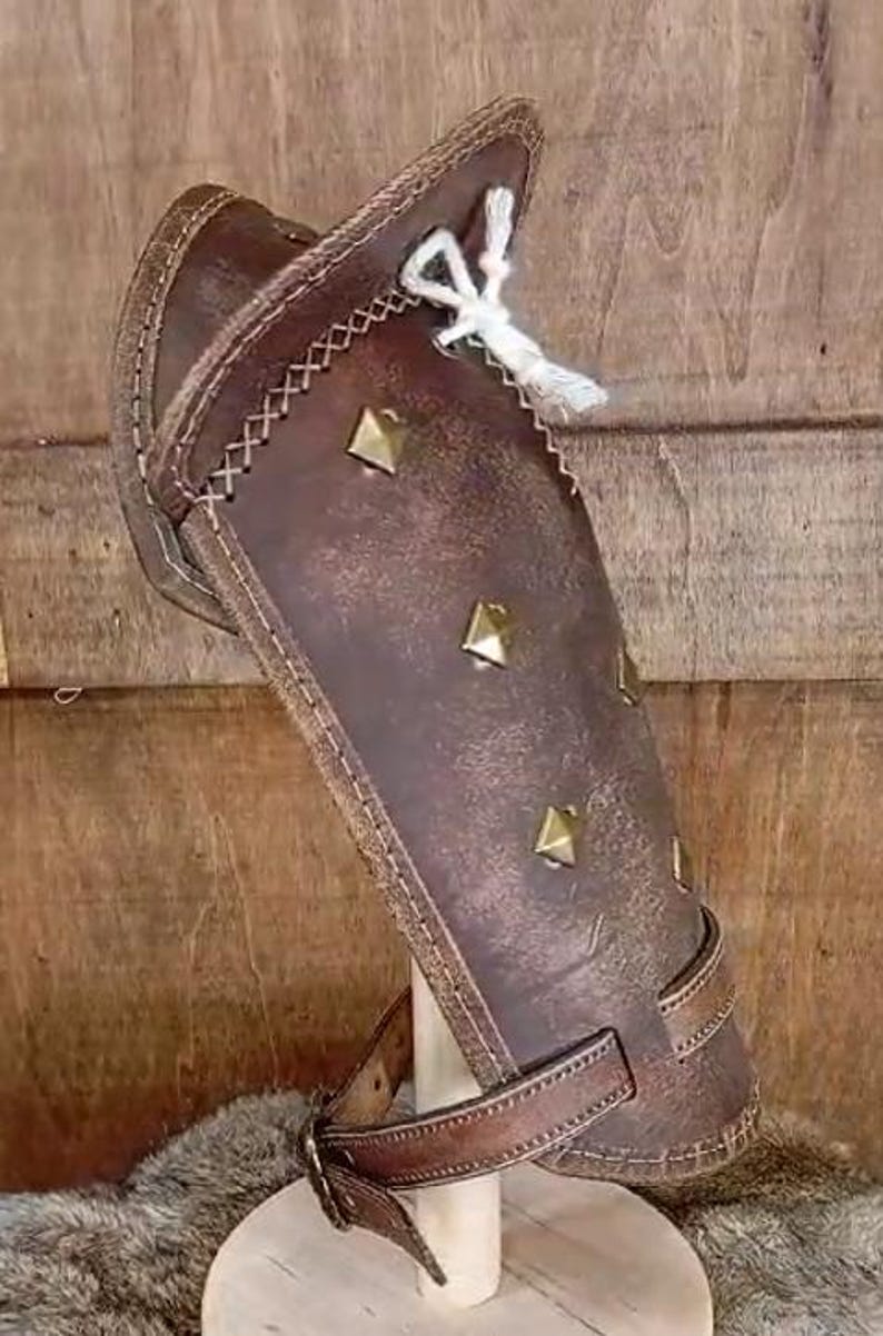 May include: Brown leather arm guard with decorative stitching and metal studs. The guard has a strap with a buckle for fastening.