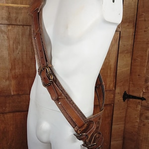 May include: A brown leather shoulder harness featuring intricate embossed border patterns, multiple adjustable straps, and antique brass-toned metal buckles. The harness is worn diagonally across the torso, with additional smaller straps and buckles attached.