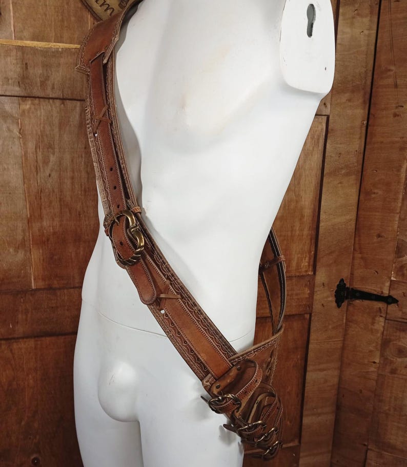 May include: A brown leather shoulder harness featuring intricate embossed border patterns, multiple adjustable straps, and antique brass-toned metal buckles. The harness is worn diagonally across the torso, with additional smaller straps and buckles attached.