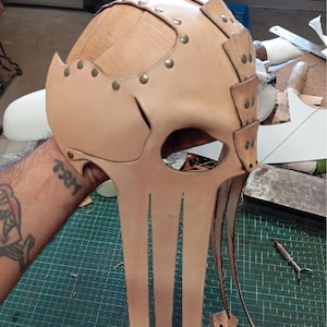 May include: A light tan leather mask featuring a segmented dome construction with metallic rivets. The design includes a large eye opening and multiple long, dangling leather strips extending downward from the base of the face piece.