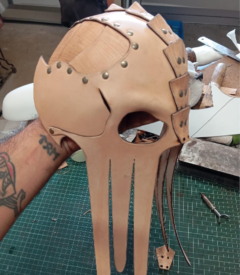 May include: A light tan leather mask featuring a segmented dome construction with metallic rivets. The design includes a large eye opening and multiple long, dangling leather strips extending downward from the base of the face piece.