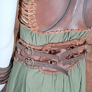May include: A brown leather corset with side lacing and gold-toned eyelets, worn over a green dress. The corset features a wide waist belt with two horizontal straps, antique-style metal buckles, and decorative brown fur-like trim along the edges.