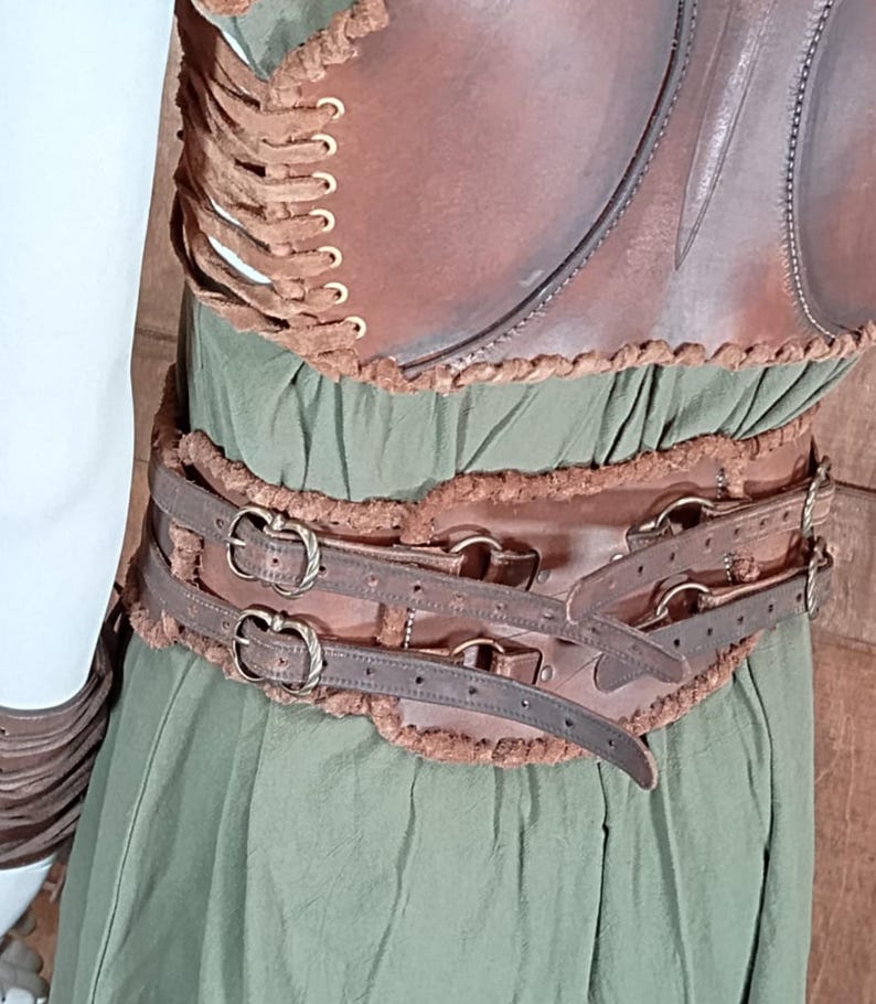 May include: A brown leather corset with side lacing and gold-toned eyelets, worn over a green dress. The corset features a wide waist belt with two horizontal straps, antique-style metal buckles, and decorative brown fur-like trim along the edges.