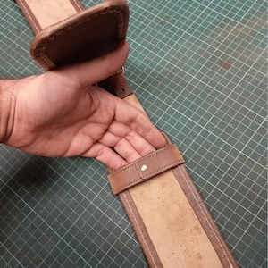 May include: A brown leather sword belt with a padded top and a rectangular sheath. The belt features a buckle and a strap with a metal stud. The leather has a worn appearance.