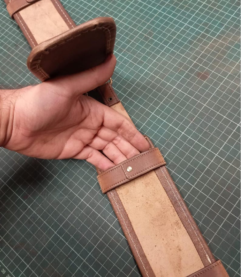 May include: A brown leather sword belt with a padded top and a rectangular sheath. The belt features a buckle and a strap with a metal stud. The leather has a worn appearance.