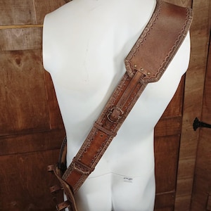 May include: A brown leather shoulder strap featuring intricate embossed scalloped edges and decorative stitching. The strap includes a metal buckle for adjustment and a wide shoulder pad section, secured with metal rivets along the length of the strap.