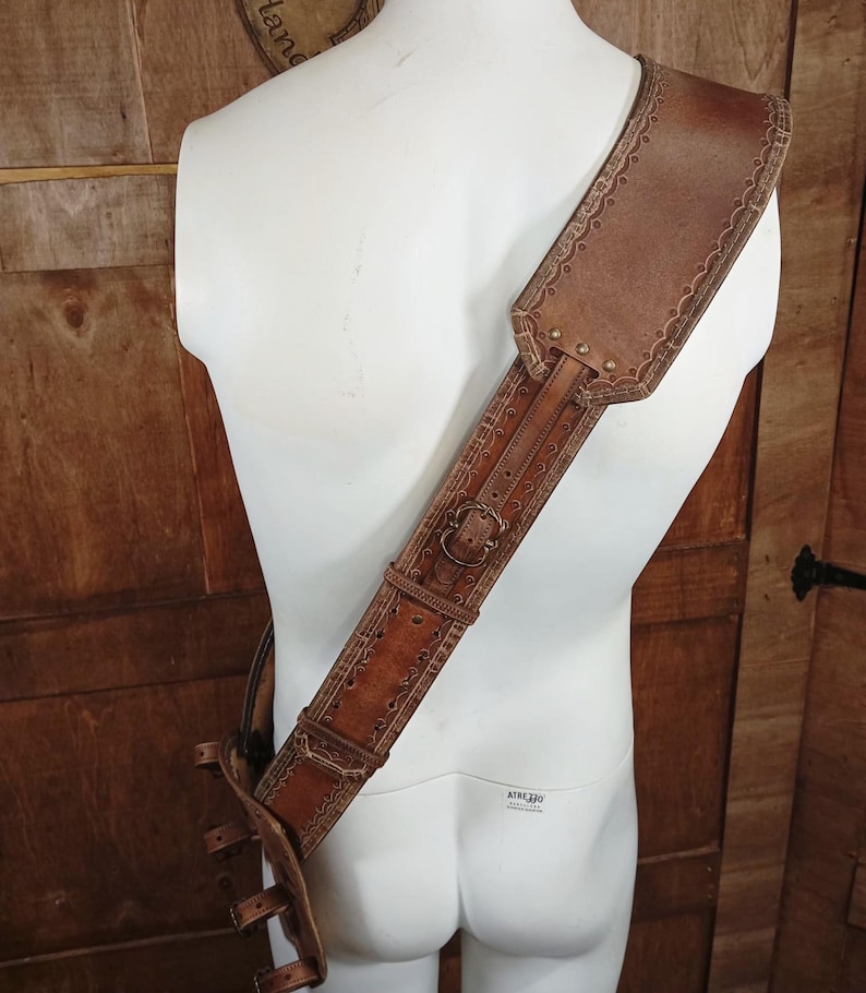 May include: A brown leather shoulder strap featuring intricate embossed scalloped edges and decorative stitching. The strap includes a metal buckle for adjustment and a wide shoulder pad section, secured with metal rivets along the length of the strap.