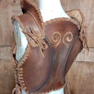 May include: A brown leather bodice featuring a dark brown finish, intricate embossed spiral patterns on the back, and decorative braided leather trim along the edges. The garment includes side lacing with brass-toned eyelets and leather ties.