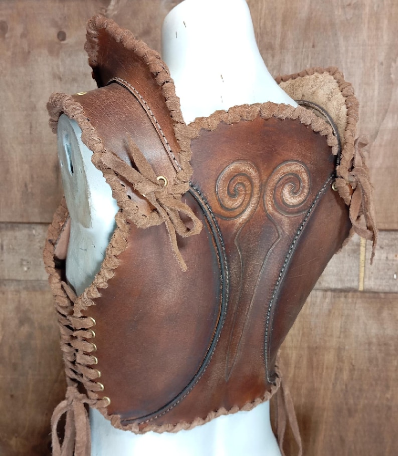May include: A brown leather bodice featuring a dark brown finish, intricate embossed spiral patterns on the back, and decorative braided leather trim along the edges. The garment includes side lacing with brass-toned eyelets and leather ties.