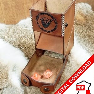 Puede incluir: Una torre de dados de cuero marrón con una bandeja, con un diseño cosido y un emblema circular con una ilustración de un goblin. Dos dados naranjas descansan en la bandeja. El texto "Friki Tournament" es visible. Un gráfico de "Descarga digital PDF" está en la esquina.